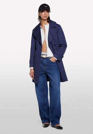 Marinefarbener Trenchcoat mit breiten Revers, kombiniert mit einem weißen Oberteil und blauen Schlagjeans. Eine schwarze Kappe, die das Outfit ergänzt, trägt ein Logo.