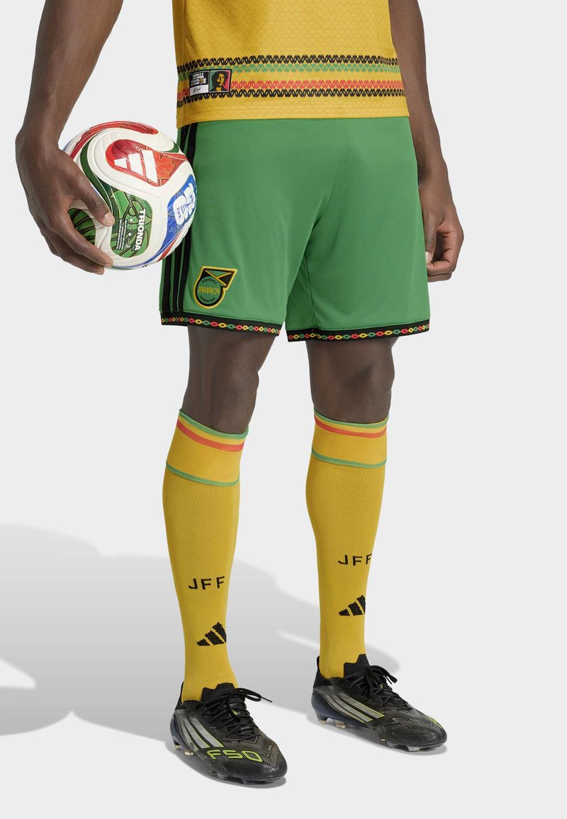 Mannelijke voetballer in geel shirt, groene broek met Jamaica-embleem, gele sokken met "JFF," zwarte voetbalschoenen, en hij houdt een kleurrijke voetbal vast.