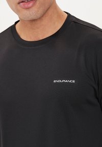 Schwarzes T-Shirt mit kurzen Ärmeln aus glattem Stoff, mit rundem Halsausschnitt und kleinem weißen "ENDURANCE"-Logo auf der Brust.