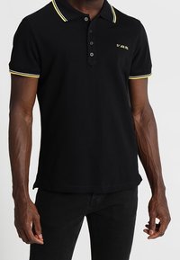 Svart polo skjorta med gul trim längs kragen och ärmarna; har tre knappar och en liten logotyp på vänster bröst.