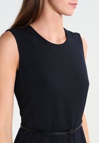 Svart ärmlös klänning med rund halsringning, som har en figurnära silhuett och ett tunt svart bälte i midjan för en skräddarsydd effekt.