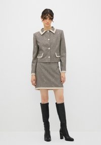 Blazer in tessuto houndstooth grigio con colletto, bottoni e gonna coordinata; abbinato a stivali neri con tacco fino al ginocchio per un contrasto.