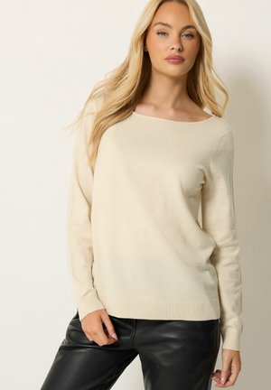 Kvinde med langt blondt hår iført en cremefarvet sweater og sorte læderbukser, stående mod en enkel lys baggrund.