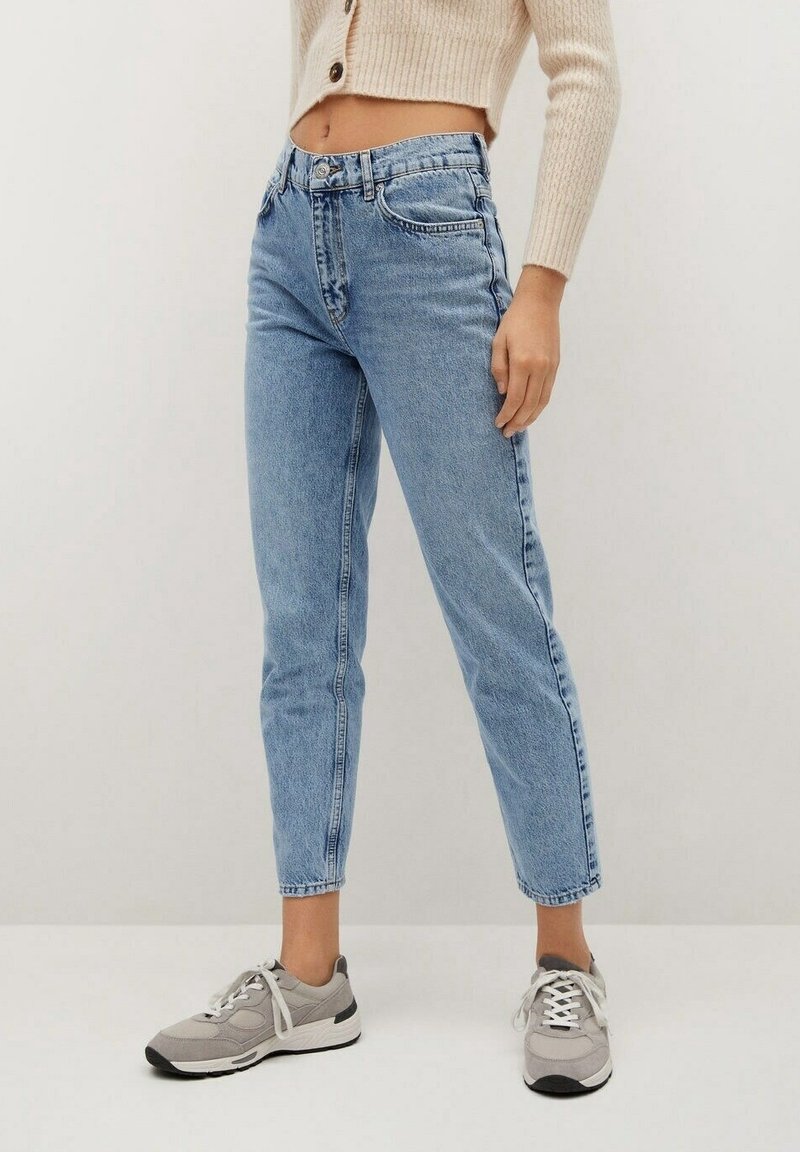 Woman wearing light blue high-waist mom jeans, beige cropped cardigan, and grey sneakers standing against plain background.