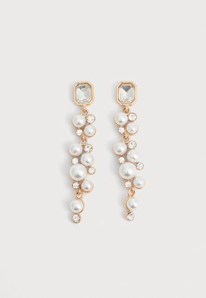 Paire de boucles d’oreilles pendantes dorées, ornées de pierres transparentes rectangulaires, de perles blanches suspendues et de petits cristaux transparents sur un fond clair.