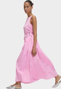 Robe maxi rose avec un design sans manches, un tissu texturé et une taille ceinturée avec des accents métalliques. Portée avec des chaussures à brides arrière argentées.