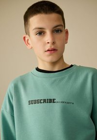 Mintgrøn sweatshirt lavet af blødt bomuld, med sort tekst "SUBSCRIBE LDN 51.5229° N. 0.0777° W" på forsiden. Klassisk crew neck design.