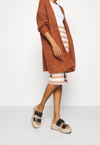 Roestkleurige gebreide cardigan met losse mouwen, witte top, gestreepte rok in beige en wit, en zwarte platform sandalen met gespsluiting.