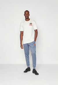 Witte korte mouw t-shirt met rood Adidas Football-logo, gecombineerd met lichtblauwe joggingbroek met zwarte accenten en zwarte sportschoenen.