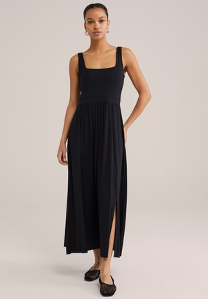 Zwart maxi-jurk met een vierkante halslijn, aansluitend lijfje en vloeiende rok met zijsplits. Model draagt zwarte mesh instappers.