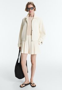 Femme portant une veste crème oversize, un short plissé assorti, un haut beige, des lunettes noires, des tongs et tenant un sac cabas noir.