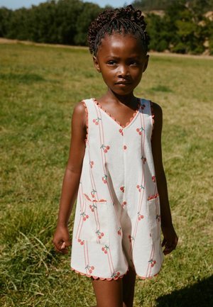 Młoda dziewczynka z warkoczami, ubrana w białe, bezrękawnikowe kombinezon z nadrukiem wiśni, stoi na trawiastej polanie w słoneczny dzień.