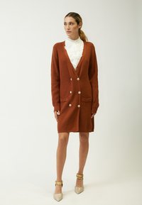 Cardigan de color óxido con escote en V, mangas largas, dos bolsillos frontales y botones grandes. Llevado sobre una blusa blanca con volantes, combinado con tacones brillantes.