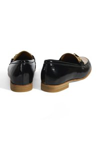 Zwarte gepatineerde leren instappers hebben een goudkleurige accent aan de voorkant, een glad bovendeel en een tan rubberen zool met een lage hak.