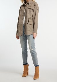 Olivgrüne Utility-Jacke mit mehreren Fronttaschen, Knopfverschluss und entspannter Passform, kombiniert mit hellblauen, ausgewaschenen Jeans und beigefarbenen Stiefeletten.