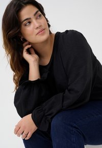 Black, textured top with balloon sleeves and ruffle detail, paired with dark blue jeans. Model's hand rests on knee, wearing minimal rings.