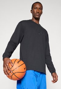 Schwarzes Langarmshirt mit einem strukturierten Logo, kombiniert mit blauen Shorts. Die Person hält einen orangefarbenen Basketball. Schlichter Hintergrund.