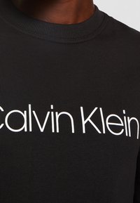 Sudadera de algodón negra con cuello redondo acanalado y un prominente logo blanco de "Calvin Klein" impreso en la parte frontal.