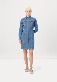 Denim košulja haljina s dugim rukavima, gumbima sprijeda, dva džepa na prsima i dekorativnim zakovicama na ramenu; svijetloplava boja i do koljena.