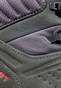 Grønn fottur sko med en nubuck-overdel og teksturerte mørke tekstildetaljer, detaljert søm og en kontrasterende logo i oransje.