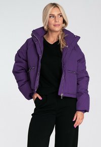 Fioletowa krótka kurtka puchowa z poziomym pikowaniem, zapinanymi kieszeniami oraz dużym kapturem, noszona na czarnym topie. Gładkie wykończenie.