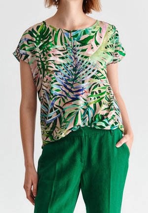 Femme portant un chemisier à manches courtes avec un imprimé de feuilles tropicales vertes et bleues, rentré dans un pantalon vert taille haute, une main dans la poche.