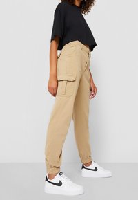 Beiga cargobyxor med elastiska muddarn och sidofickor, ihop med en kort svart t-shirt och vita sneakers med svarta detaljer.