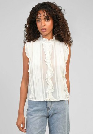 Femme aux cheveux bouclés portant un chemisier blanc sans manches à volants et un jean bleu clair avec des détails subtils scintillants sur un fond uni.