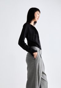 Musta pitkähihainen paita, jossa on kerätty pääntie, yhdistettynä vaaleanharmaisiin raidoitettuihin housuihin. Housuissa on taskut ja nappikiinnitys.