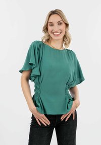 Blusa teal con maniche corte e arricciate e dettaglio laterale raccolto. Abbinata a jeans neri. Texture liscia, design casual.