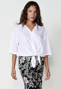 Blusa bianca leggera con un davanti annodato, patta dei bottoni, e maniche scese, abbinata a pantaloni neri con motivo floreale.