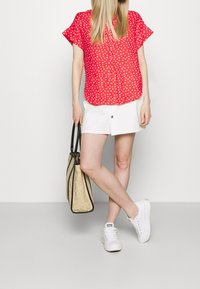 Czarna bluzka w czerwone kropki z krótkim rękawem w połączeniu z białą mini spódnicą i białymi trampkami, trzymając złotą torbę ze słomy.