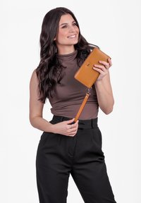 Brown leather wallet with detachable strap, smooth texture, rectangular shape, and a subtle logo. Held by a person wearing a sleeveless top.