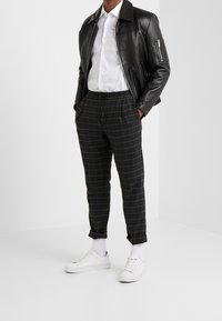 Chaqueta de cuero negra sobre una camisa blanca abotonada, combinada con pantalones de cuadros grises y zapatillas blancas, con cintura ajustable con cordón y puños enrollados.