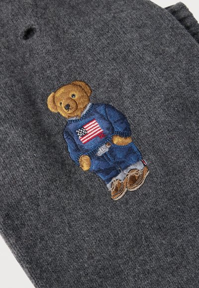 Ours en peluche marron brodé sur tissu gris, portant un jean bleu, un pull bleu avec un drapeau américain et des chaussures marron.