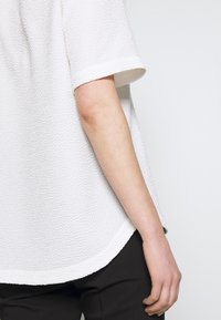 Vitt texturerat kortärmat linne med rundad nederkant. Tyget har ett lätt upphöjt mönster. En hand syns i kanten, vilande naturligt.