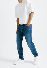 Vaqueros de mezclilla azul con pierna recta, combinados con una camiseta blanca de manga corta y zapatillas blancas. Los vaqueros cuentan con bolsillos clásicos y costuras tradicionales.