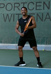 Cămașă fără mâneci neagră cu logo-ul "BOSS", pantaloni scurți negri și adidași negri. Ține o rachetă de tenis portocalie pe un teren verde cu un fundal de plasă.