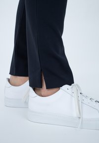 Pantaloni sportivi neri con spacchi laterali, abbinati a sneakers bianche in pelle con punta tonda e lacci piatti. Design minimalista e testurizzato.