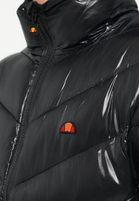 Juoda pūkinė striukė blizgiu paviršiumi, pasižyminti kūgine siūlelių raštu. Turi užtrauktuką priekyje ir mažą spalvotą logotipą krūtinės srityje.