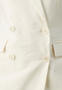 Blazer sur mesure beige clair avec une texture lisse, deux poches à rabat et six boutons ronds de couleur crème sur le devant.