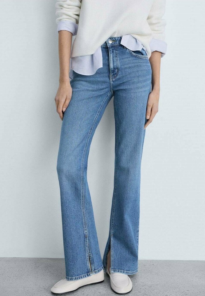 Jeans de mezclilla azul claro acampanados con aberturas laterales en el dobladillo, de cintura alta y costuras visibles. Combinados con un suéter blanco de canalé.