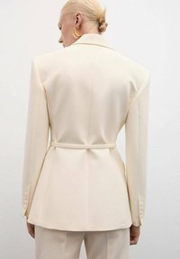 Krem blazer z oblikovanimi rameni, prilegajočim pasom in diskretnim peplumom. Ima pas na hrbtni strani in gumbne detajle na rokavih.