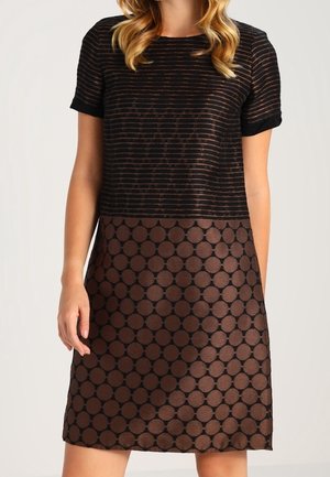 Mujer con un vestido de manga corta con rayas horizontales negras en la parte superior y grandes lunares marrones en la falda.
