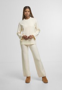 Maglione lavorato a maglia color crema con polsini a coste, abbinato a pantaloni larghi coordinati. Le calzature presentano stivali alla caviglia in suede marrone con suola spessa.