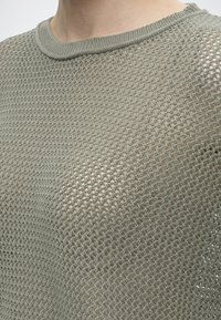 Gros plan d'une personne portant un pull en maille légère gris clair, tissé de façon lâche, avec un col rond, laissant voir la peau en dessous.