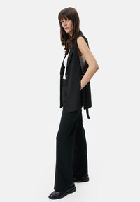 Schwarze ärmellose Blazer, kombiniert mit einem enganliegenden weißen Top und weit geschnittenen schwarzen Hosen, aus glattem Stoff und maßgeschneidertem Design.