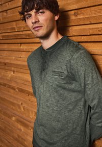 Dlouhý rukáv, tmavě zelená henley košile s texturovaným povrchem, se třemi knoflíky a logem na hrudi. Umístěná na dřevěném pozadí.