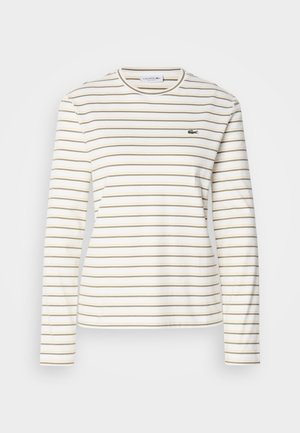 Bijela košulja dugih rukava s tankim bež horizontalnim prugama; ima okrugli izrez i Lacoste logo na lijevoj strani prsiju.