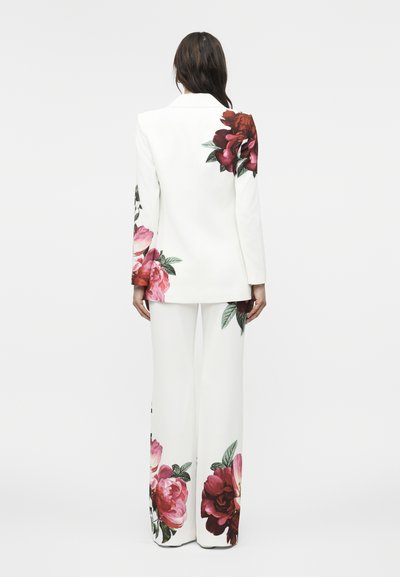 Γυναίκα με λευκό κοστούμι με μεγάλα κόκκινα και ροζ λουλουδένια σχέδια στον ώμο του σακακιού, το μανίκι και τα μπατζάκια, στεκόμενη με πλάτη προς το μέρος μας.